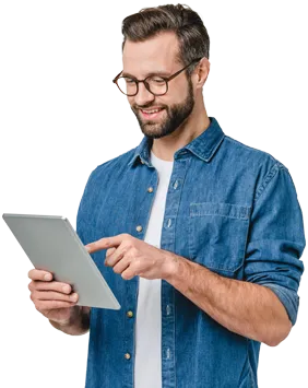 Smiling bearded man wearing glasses and a denim shirt holding and tapping a tablet device.